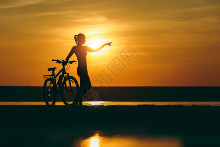 一个穿着西装的运动型女孩的剪影站在水中的一辆自行车附近图片