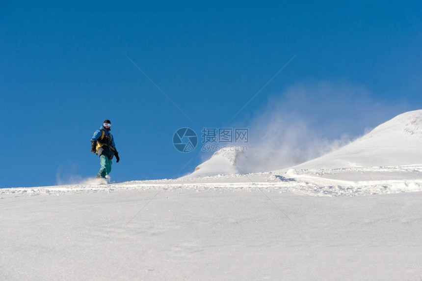 穿着滑雪面罩和在雪覆盖的山坡上用背着雪粉对着蓝天而留图片