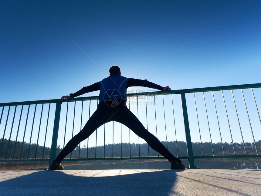 穿着高大黑色紧身裤的晨跑者使身体在桥路上伸展在湖桥上光滑的混凝土地面上进行户外锻炼太阳勾图片