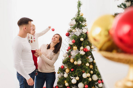 快乐年轻开朗的父母带着可爱的小儿子爸肩上的小男孩在家里的灯室里用玩具球装饰树圣诞好心情家庭爱情和假背景图片
