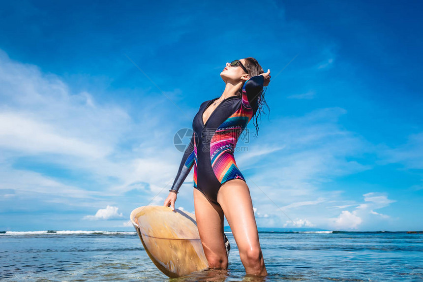 迷人的年轻女子穿着潜水服和太阳镜在印尼巴厘岛努沙杜阿海滩摆着冲浪板图片