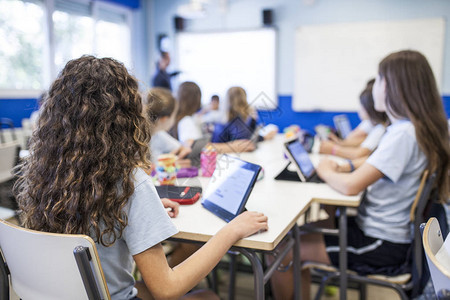 女孩棕色卷发研究在课堂上与她的平板电脑旁边的同学而他们听老师图片
