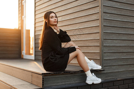时尚女人穿着时髦的衣服穿着运动鞋坐在日落的木墙附近图片