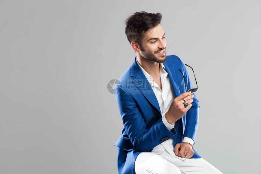 微笑智能休闲男子的肖像拿着他的太阳镜看一边而坐在浅灰色背景图片