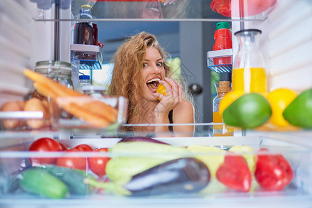 女人一边咬水果一边站在装满杂货的冰箱前从冰箱里拍的照片图片