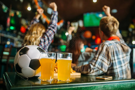 校庆球和啤酒在体育酒吧的桌子上足球迷在后台电视广播观看游戏概念背景