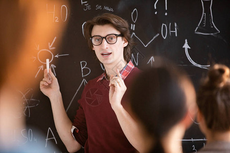 年轻有自信的眼镜老师站在黑板上拿着配方程式向学生们图片