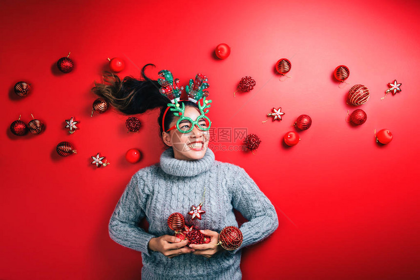 轻女子穿着温暖的毛衣与道具球红色与圣诞饰品在假日闪耀红色背景图片