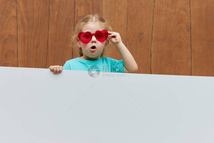 小微笑的儿童女孩心形眼镜站在木制背景的白色空白面板后面搞笑的脸从横幅后面窥视图片