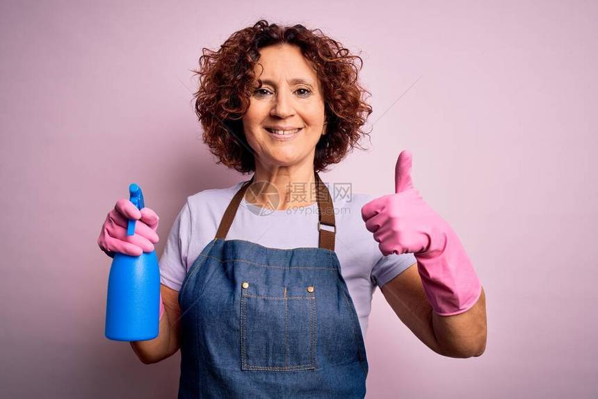 中年卷发女清洁做家务时穿着围裙和手套使用喷雾器开心地笑着做好的手势图片
