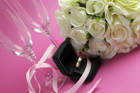 粉红色背景的白玫瑰新娘花束与一对香槟长笛眼镜和黑首饰盒中的金婚戒指图片