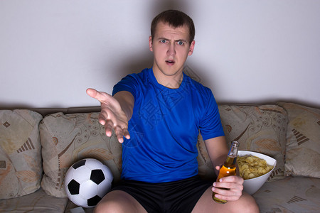 穿着制服坐在沙发上看足球用啤酒和薯片看足球的年轻激动人心图片