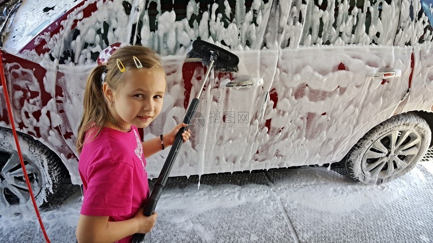 快乐的小女孩在洗车时用刷子和被肥皂覆图片