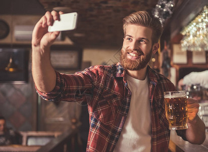 英俊的胡子帅哥在酒吧柜台喝啤酒时用智能手机做自图片
