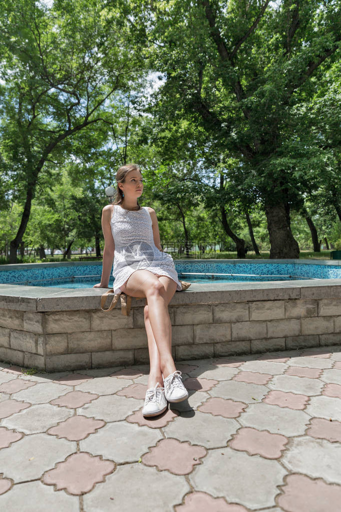 身着短白色短裙的年轻美丽的高加索女孩坐在城市公园喷泉附近的水泉边图片