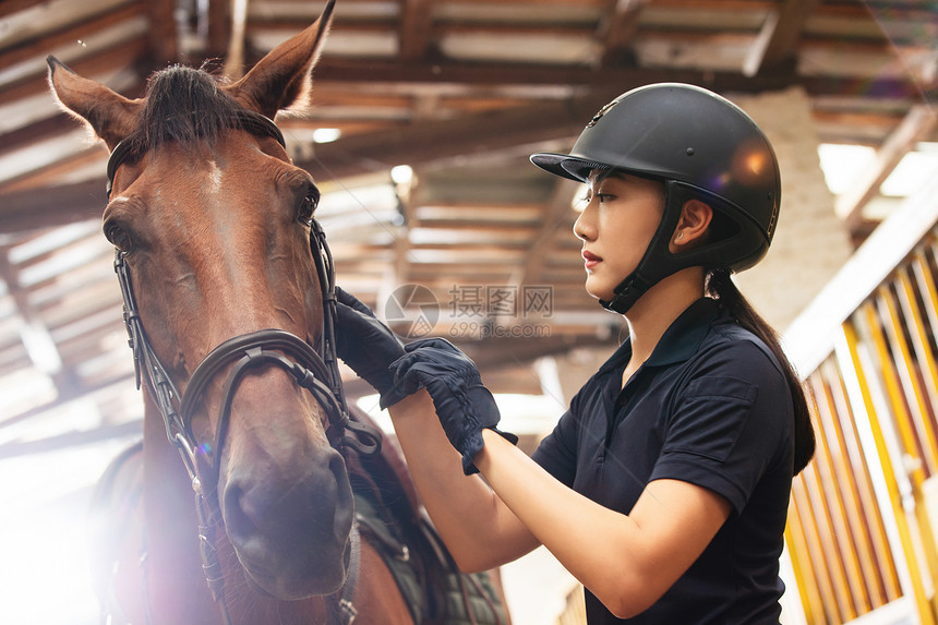 调节马的缰绳的年轻女人在马厩走廊图片