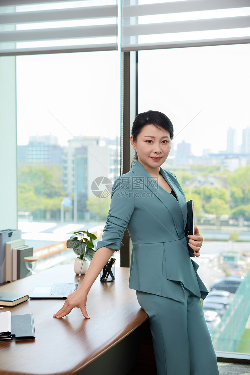女白领拿着文件夹倚靠着办公桌的形象图片