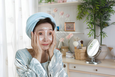 女人衰老老年女性正在使用面膜头上裹着毛巾坐在桌前对着镜子背景